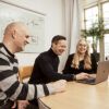 Tre kollegor ler och samarbetar kring en bärbar dator i ett ljust kontorsrum med en whiteboard i bakgrunden där det står 'SEO' och 'Ads', vilket antyder en fokusering på sökmotoroptimering och annonsering